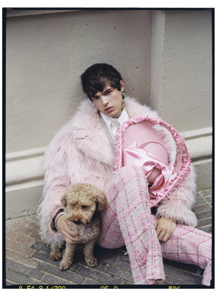 Ruslan seated with a small dog while wearing a pink faux-fur coat, tweed trousers and a ruffled hat