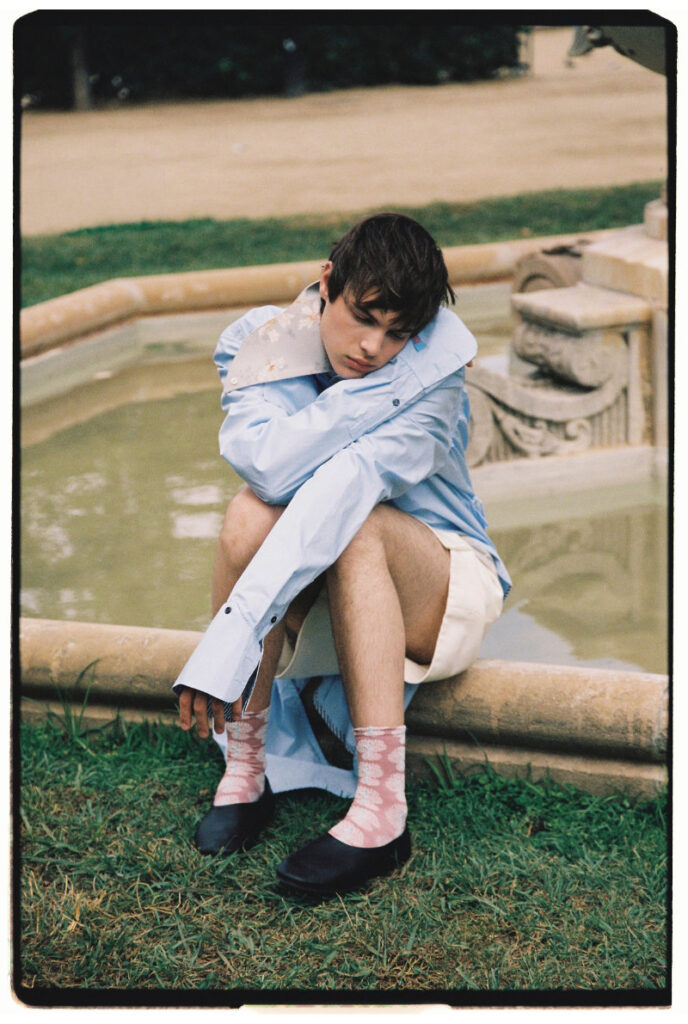 Ruslan crouching beside a fountain in an oversized blue shirt, white shorts, pink socks and black shoes