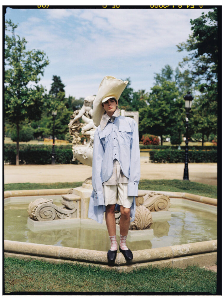Ruslan standing by a fountain in an oversized pale blue shirt, white shorts and a large cream hat