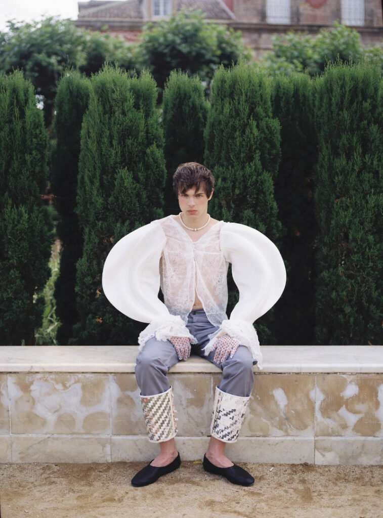Ruslan seated in a sheer white lace blouse with oversized curved sleeves and cuffed gray trousers