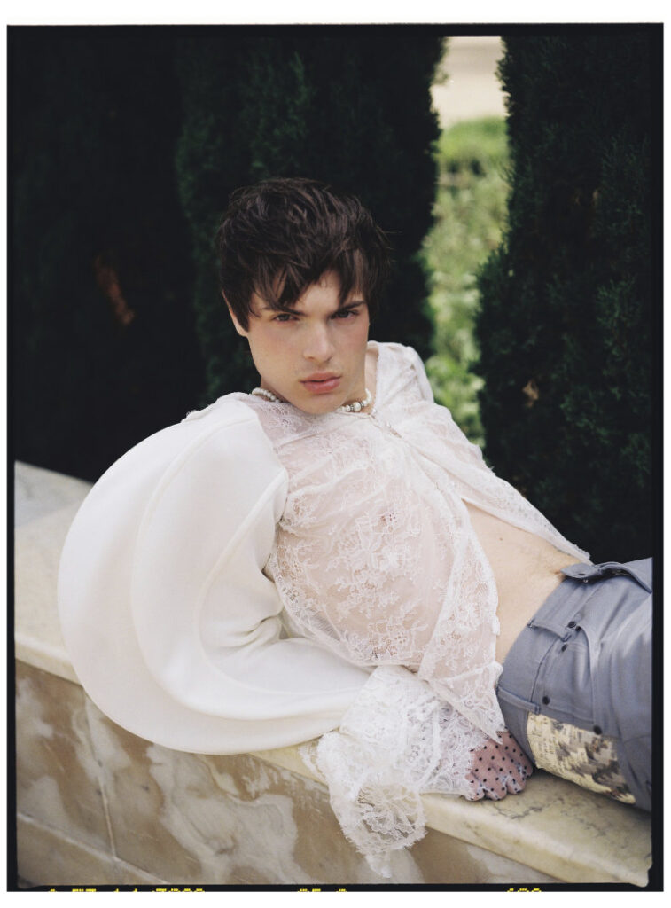 Ruslan reclining in a sheer white lace top with curved sculptural sleeves and gray trousers