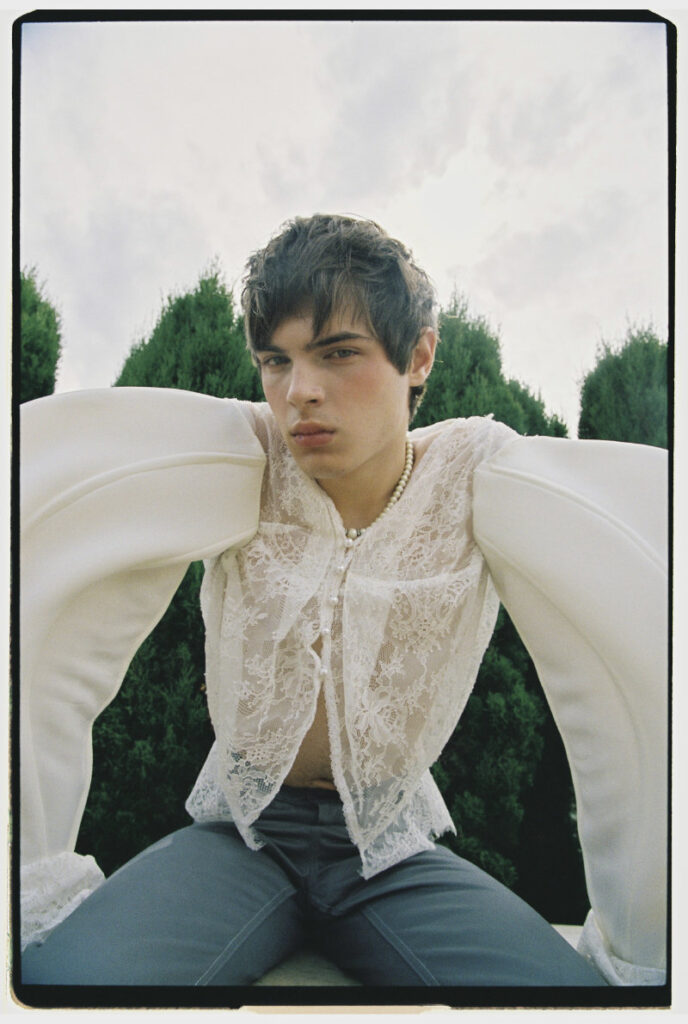 Ruslan facing the camera in a white lace top with oversized sculptural sleeves and gray trousers