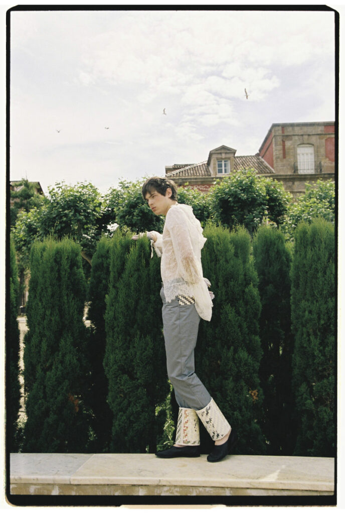 Ruslan standing beside tall garden hedges in a white lace top, gray trousers and patterned cuffs
