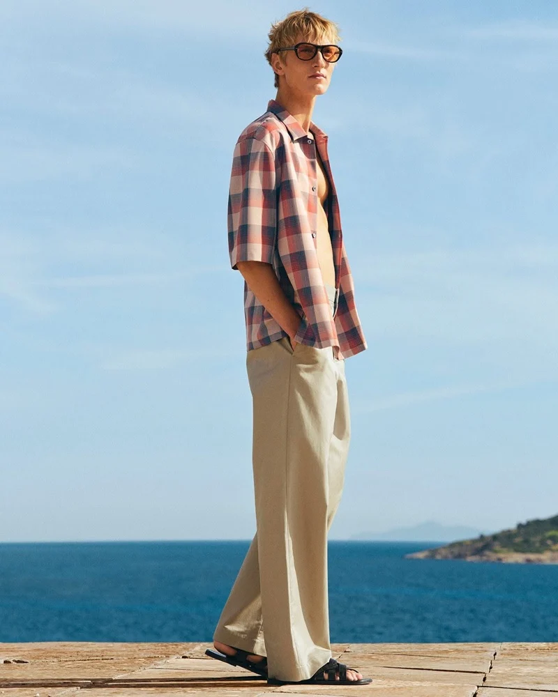 Pierrick Grégoire wearing an open red and blue check shirt with tan wide-leg trousers by the sea for COS