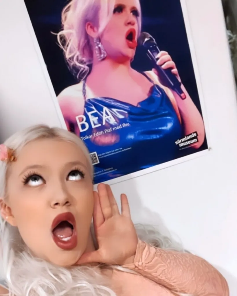 Bea MonaLisa posing beneath a poster showing her performing as Édith Piaf