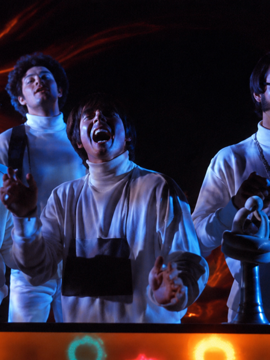 Restored still from The Monkees’ “Star Collector” video featuring the band in white outfits under blue stage lighting with a warm orange glow.