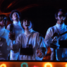 Restored still from The Monkees’ “Star Collector” video featuring the band in white outfits under blue stage lighting with a warm orange glow.
