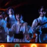 Restored still from The Monkees’ “Star Collector” video featuring the band in white outfits under blue stage lighting with a warm orange glow.