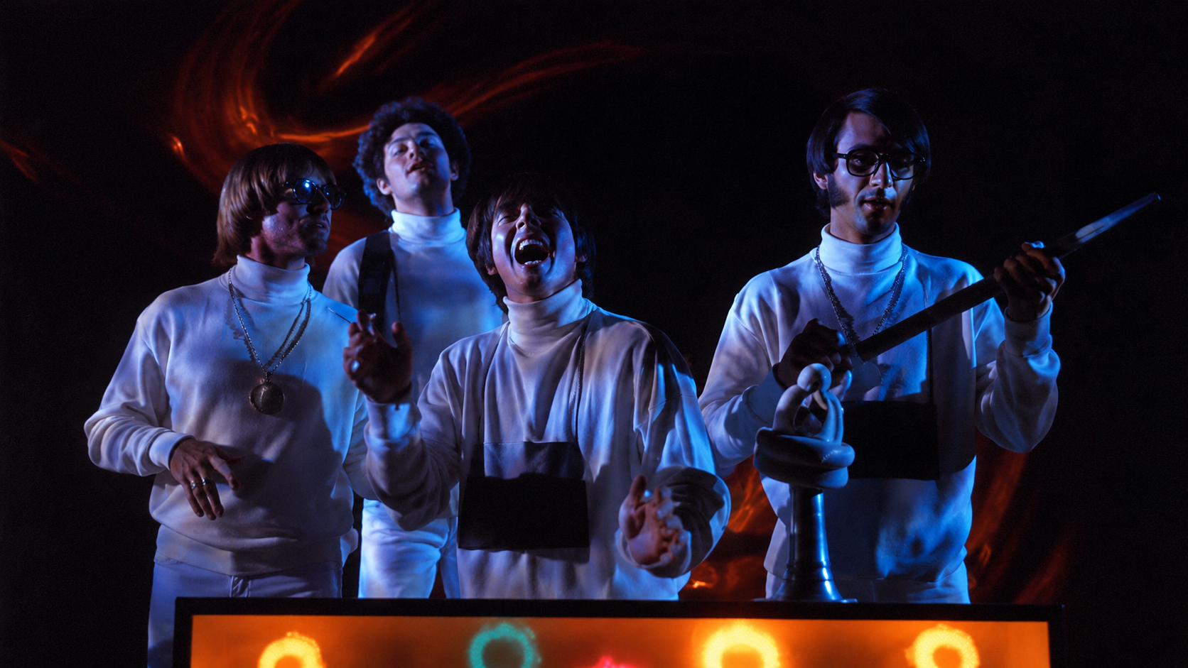Restored still from The Monkees’ “Star Collector” video featuring the band in white outfits under blue stage lighting with a warm orange glow.