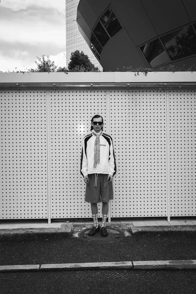 Black-and-white portrait of Anton Kügler in layered tailoring and shorts by a patterned wall