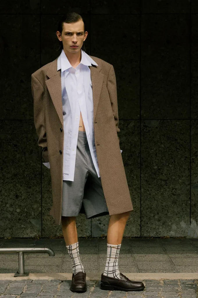 Anton Kügler in a long checked coat, open white shirt and gray shorts on a sidewalk