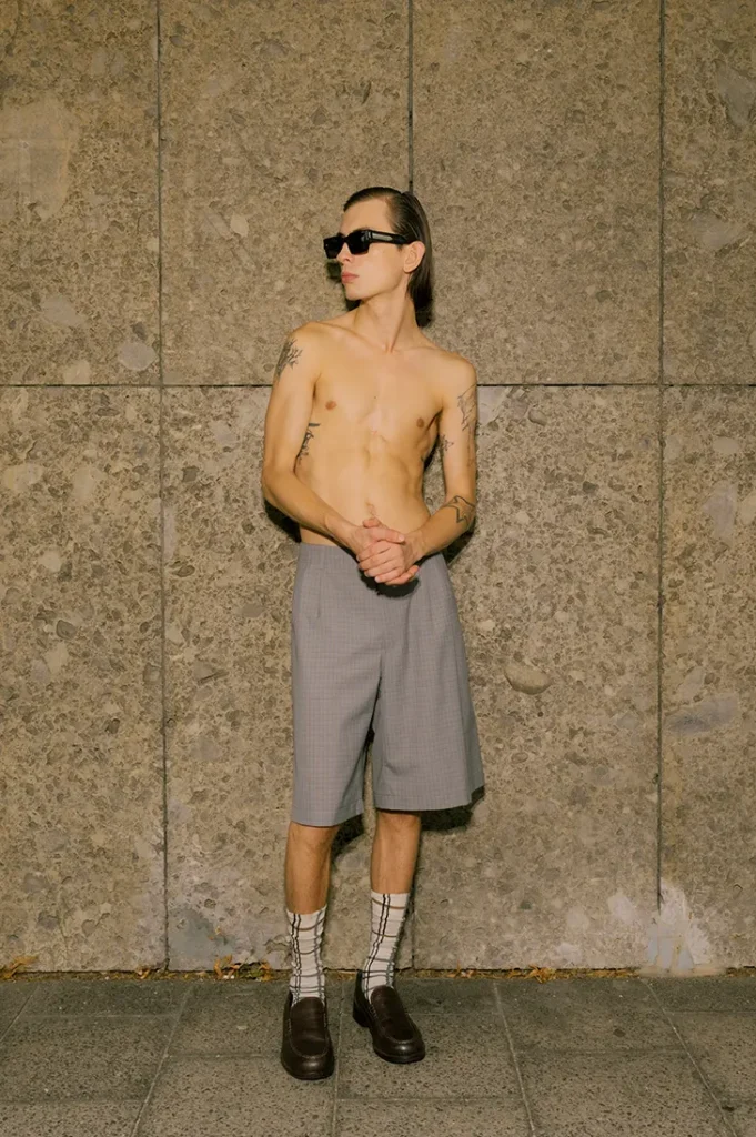 Anton Kügler shirtless in gray Bermuda shorts and sunglasses against a stone wall