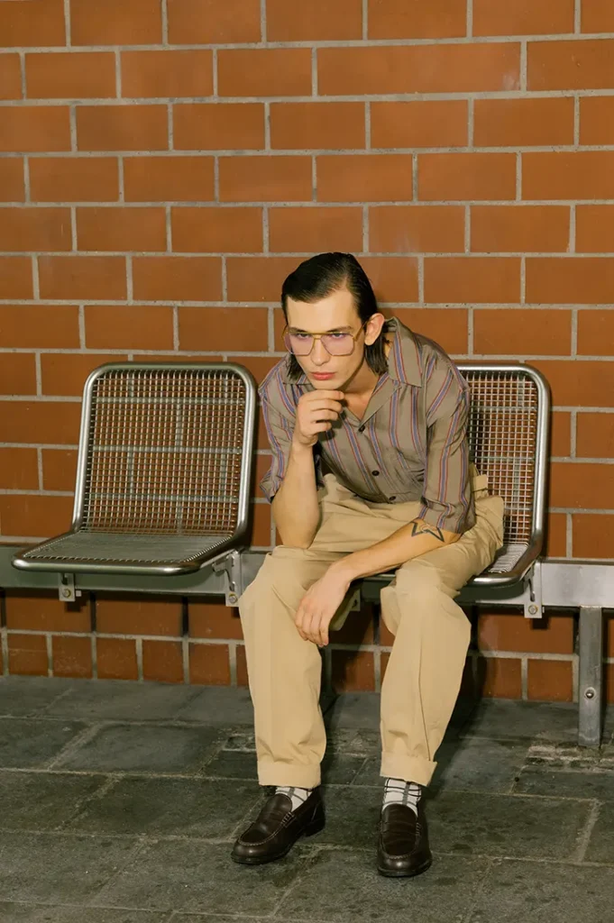 Anton Kügler seated on a metal bench in glasses, a striped shirt and beige trousers