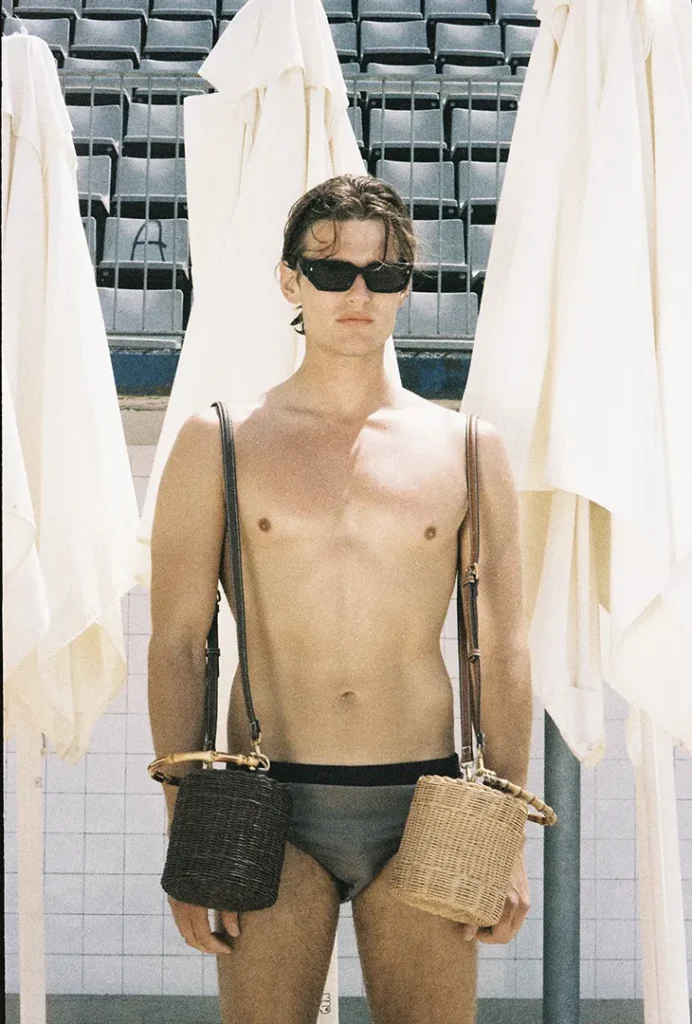 Shirtless model in sunglasses and gray briefs carrying two woven bags beside closed umbrellas