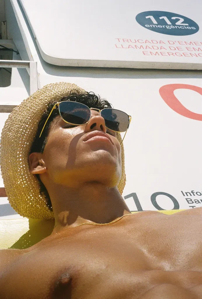 Shirtless model reclining in a straw hat and sunglasses beneath an emergency sign
