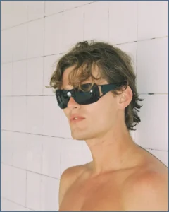 Shirtless model wearing black sunglasses against a pale tiled wall in the Nothing But Summer editorial