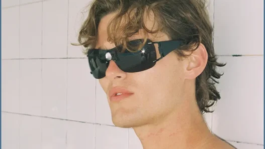 Shirtless model wearing black sunglasses against a pale tiled wall in the Nothing But Summer editorial