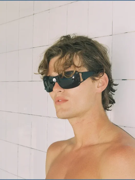 Shirtless model wearing black sunglasses against a pale tiled wall in the Nothing But Summer editorial