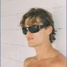 Shirtless model wearing black sunglasses against a pale tiled wall in the Nothing But Summer editorial