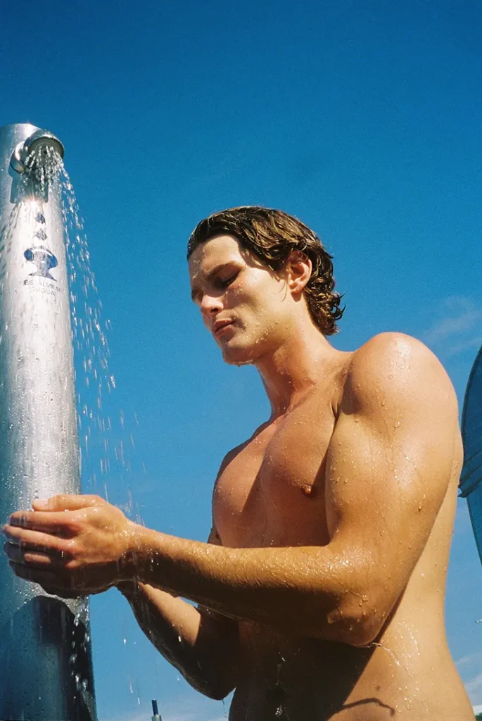 Shirtless model standing beneath an outdoor shower against a clear blue sky