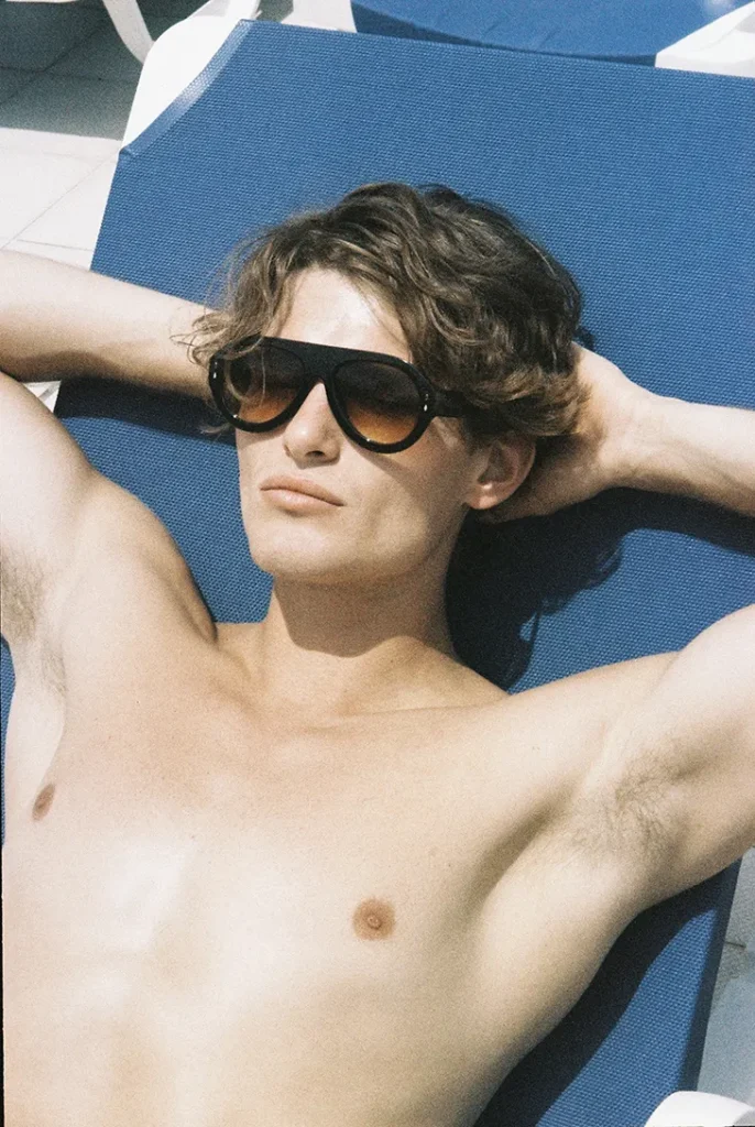 Shirtless model in black sunglasses reclining on a blue pool lounger with his hands behind his head