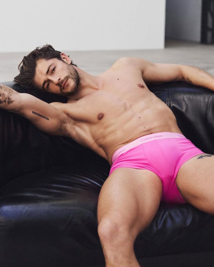 Sam Salter reclining shirtless on a black leather bench in bright pink Calvin Klein trunks