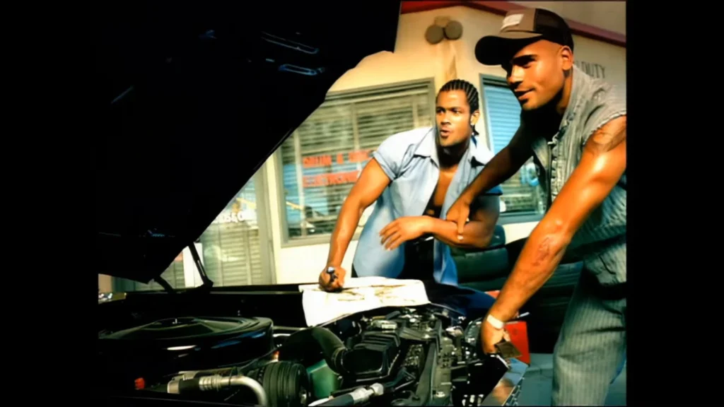 Two men look beneath the hood of a car in the “Whatever U Want” music video.