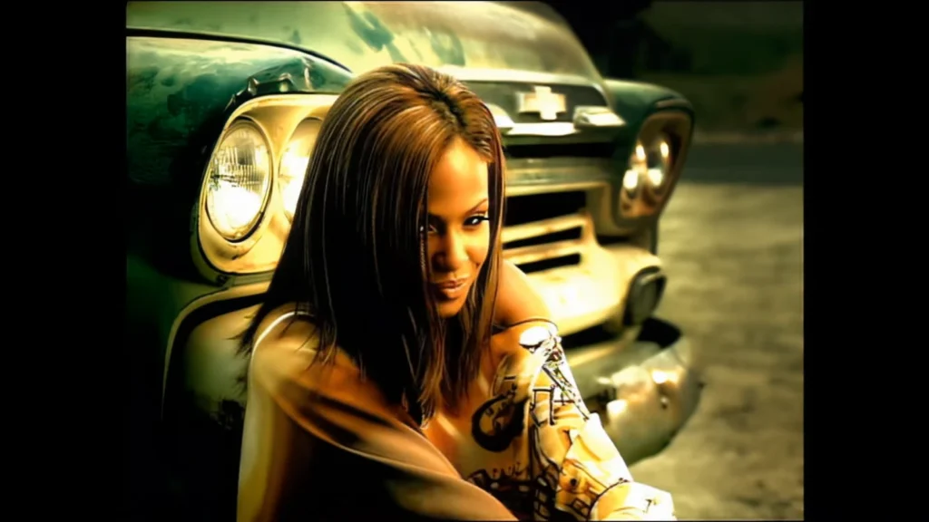 Christina Milian crouches beside the front of a car in the “Whatever U Want” music video.