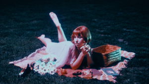 Frankie June lounges on a picnic blanket at night beside flowers and a woven basket in a vintage-inspired scene.