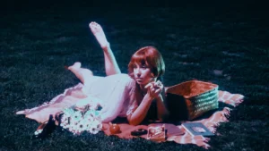 Frankie June lounges on a picnic blanket at night beside flowers and a woven basket in a vintage-inspired scene.