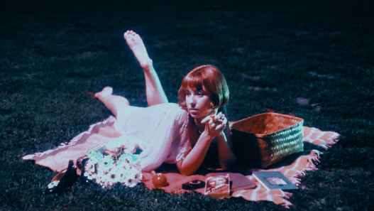 Frankie June lounges on a picnic blanket at night beside flowers and a woven basket in a vintage-inspired scene.
