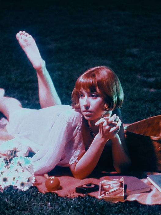 Frankie June lounges on a picnic blanket at night beside flowers and a woven basket in a vintage-inspired scene.