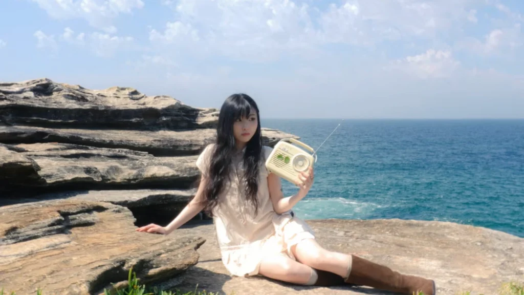 Woman holding a portable radio while seated on sunlit coastal rocks beside the ocean