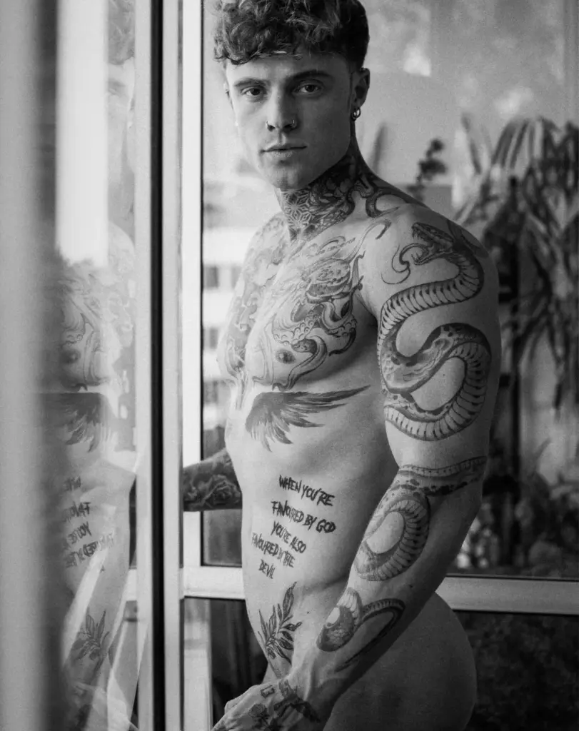 Black-and-white side portrait of Samuel Dohmen by a window, showing his tattooed shoulder and torso