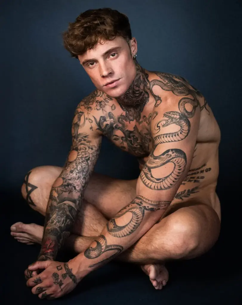 Color portrait of Samuel Dohmen crouching against a dark blue backdrop with his tattooed arms visible