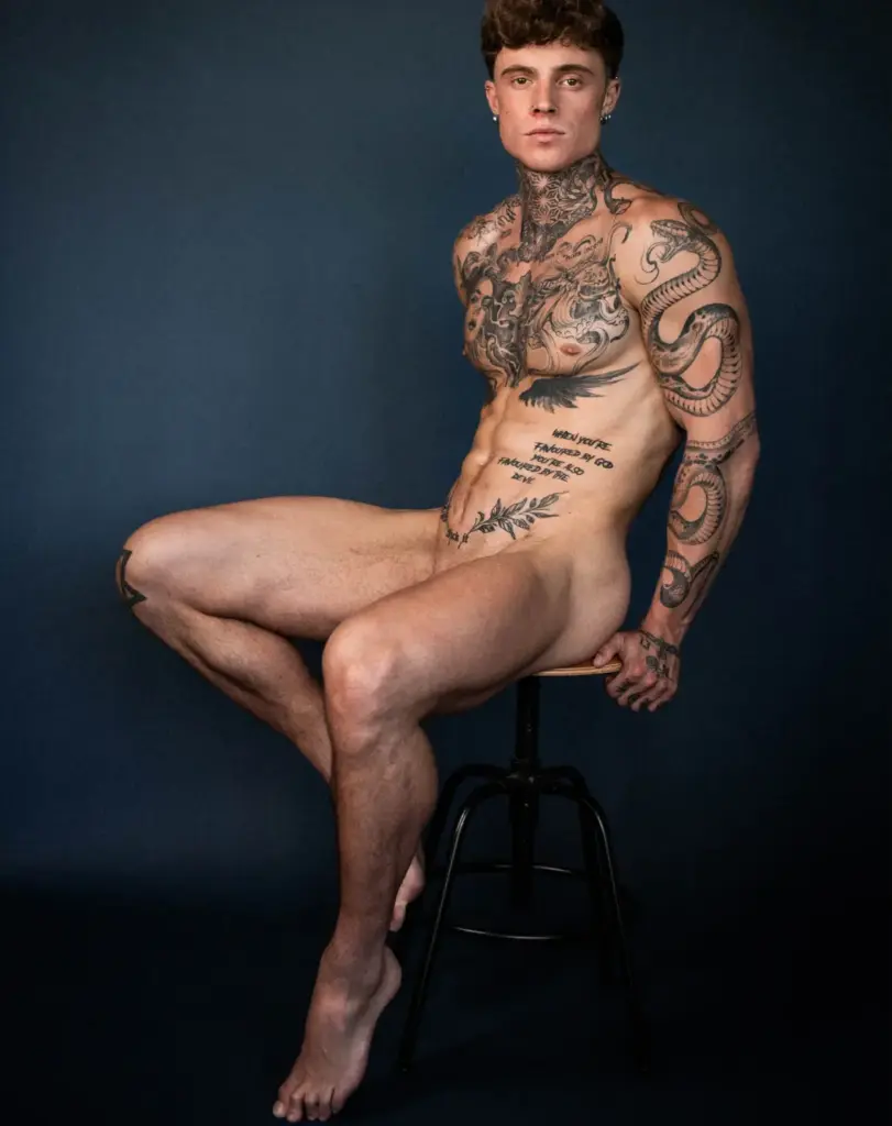 Color studio portrait of Samuel Dohmen sitting on a black stool against a blue background, showing his tattoos