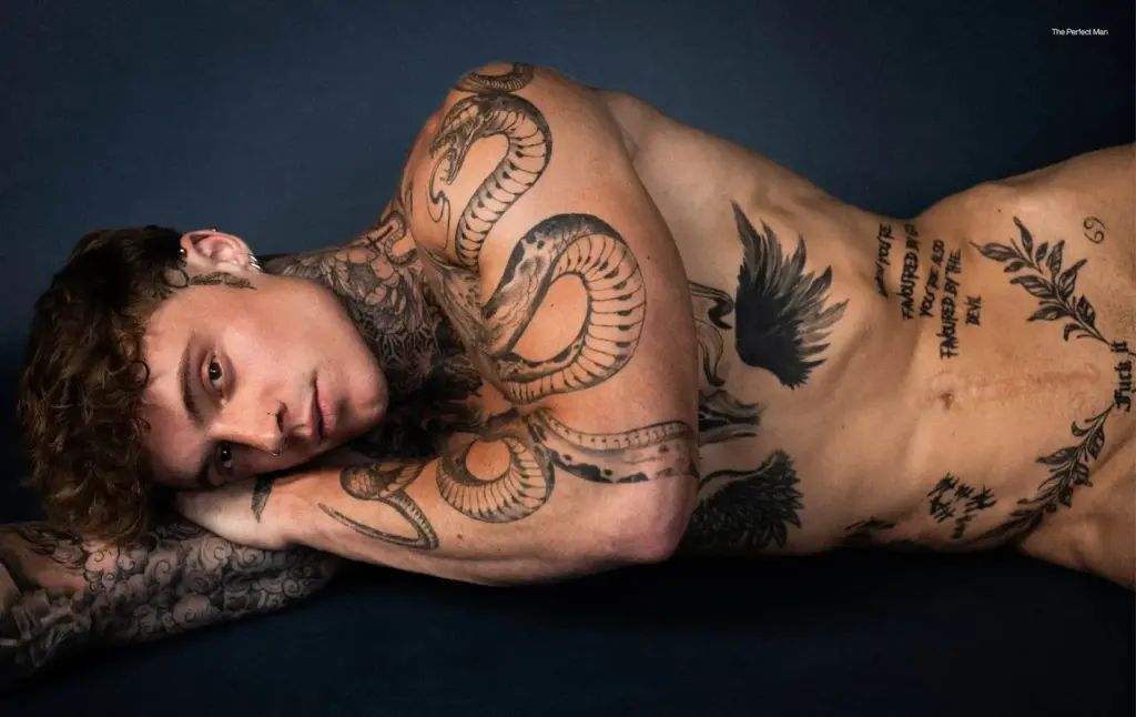 Samuel Dohmen lying on his side against a dark blue backdrop, his tattooed arms and torso visible