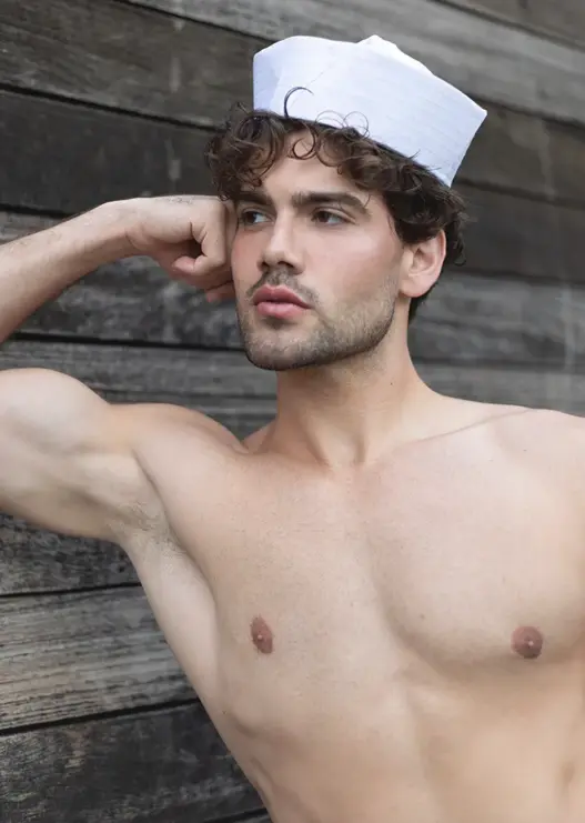 Model Amir Suarez reclining on seaside rocks in a navy blazer and white sailor cap, with geese swimming behind him, photographed by Jose Mata for MMSCENE's Autumn Afternoon on the Bay editorial.