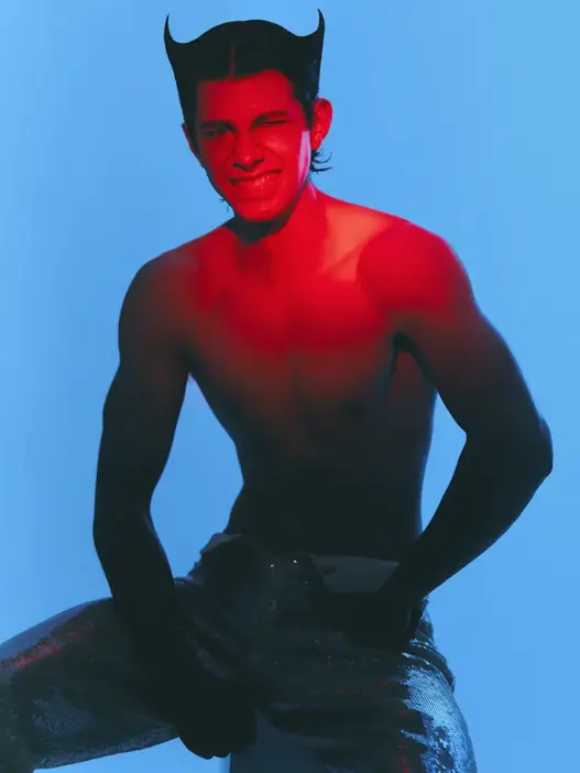 Model Ángel Sánchez poses shirtless with horn-shaped hairstyle under dramatic red lighting against a blue backdrop for Avoir de L’Allure, photographed by Robb Sb