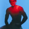 Model Ángel Sánchez poses shirtless with horn-shaped hairstyle under dramatic red lighting against a blue backdrop for Avoir de L’Allure, photographed by Robb Sb