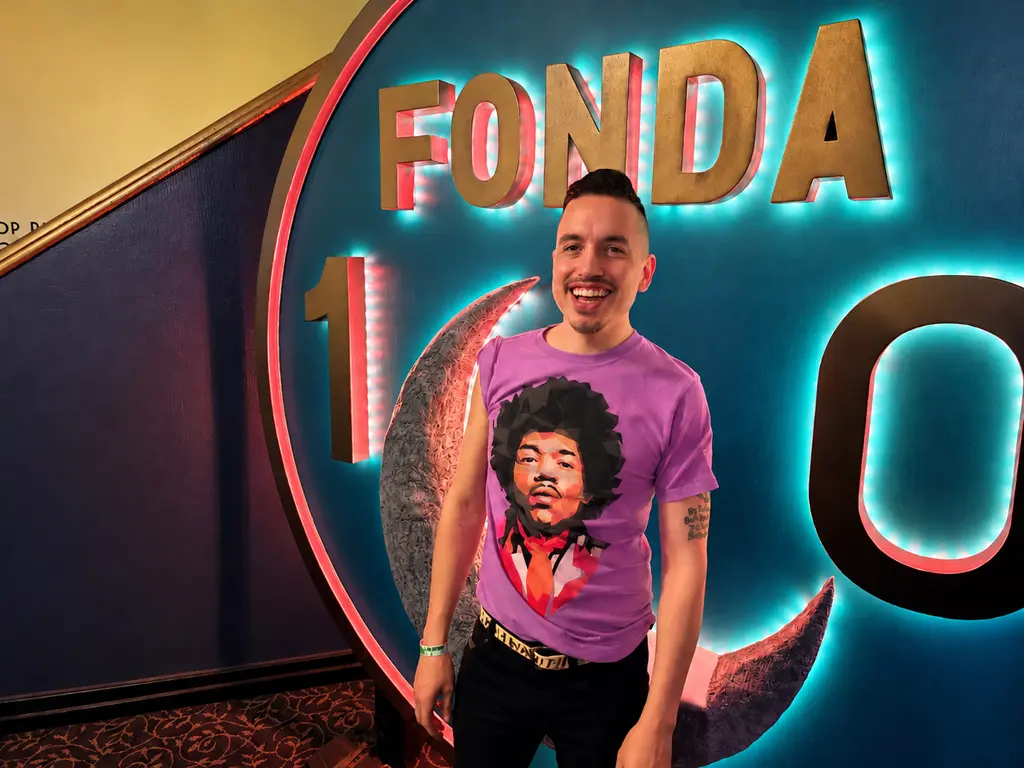 Anthony De La Cruz of INYIM Media smiling beside the illuminated Fonda Theatre 100th-anniversary sign in Hollywood