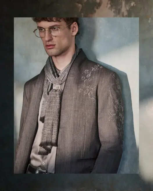 Model David Trulik wearing glasses, a textured gray blazer and layered scarf against a weathered wall in John Varvatos' Fall/Winter 2026 Wonderland campaign, photographed by Cara Johnson.