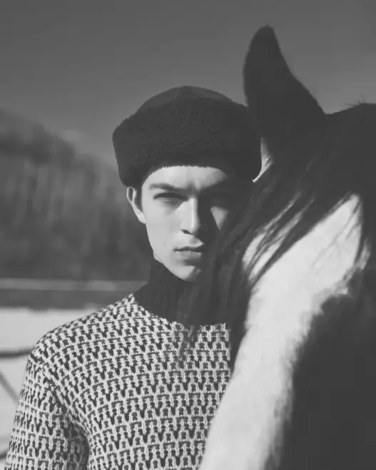 Model Just Verhoeff wearing a dark knit beanie and shearling-lined winter coat beside a horse for Loro Piana's Holiday 2026 campaign.
