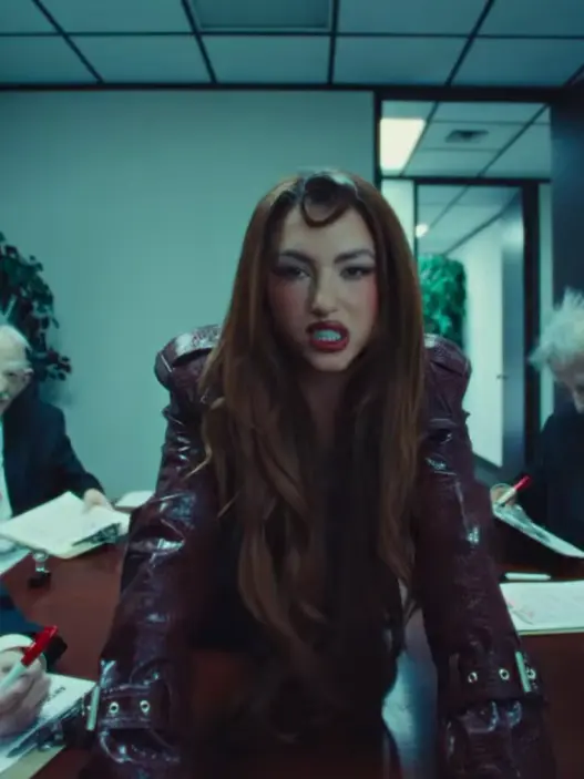 Canadian pop singer Rêve with long auburn hair, dramatic makeup and a burgundy outfit inside a blue-green office setting in her Church of Sound music video.