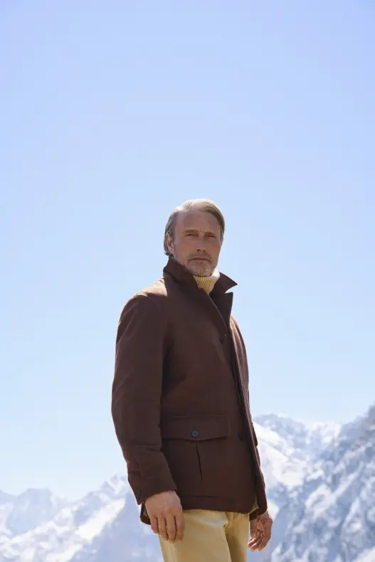 Mads Mikkelsen wearing a brown Zegna Falcon jacket over a beige turtleneck against snow-covered mountains for the Fall/Winter 2026 campaign.