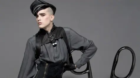 Fashion model in an avant-garde black outfit and sculptural headpiece poses beside an oversized pair of scissors against a gray studio backdrop.