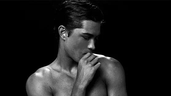 Francisco Lachowski poses shirtless in white briefs against a black background, with one hand raised near his mouth.