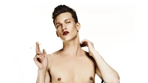 Shirtless male model with red lipstick poses against a white background in jeans, holding a black handbag and a cigarette.