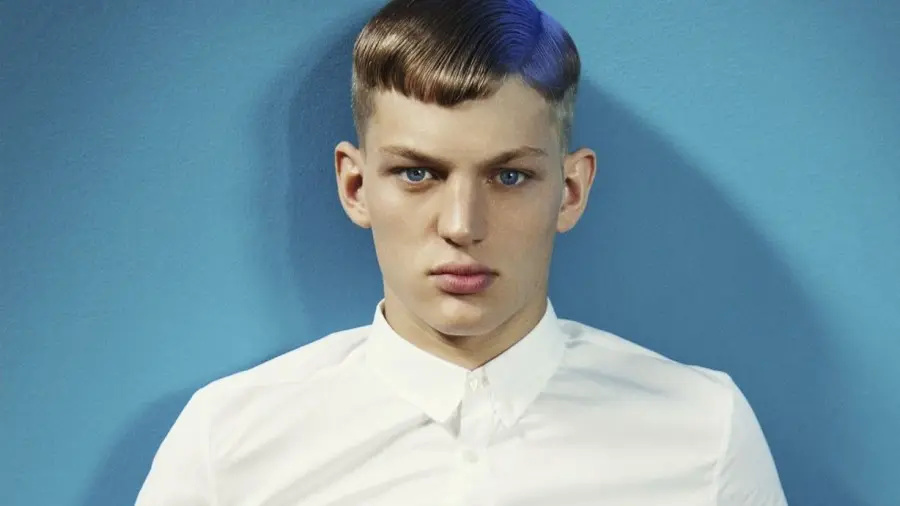 Sebastian Sauvé sits against a blue wall wearing a white button-up shirt and bright yellow shorts.