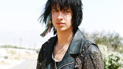 Julian Casablancas stands outdoors wearing a heavily studded black leather jacket, with shaggy dark hair and layered necklaces.